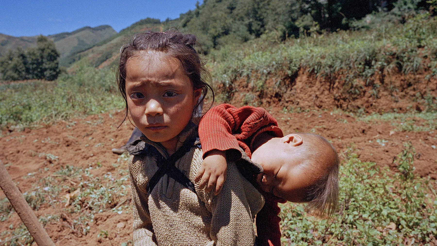 A girl carrying her younger sibling across a field in Liangshan Prefecture.