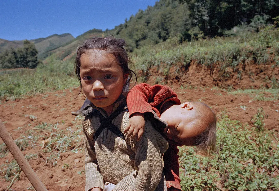 A girl carrying her younger sibling across a field, Liangshan Prefecture.